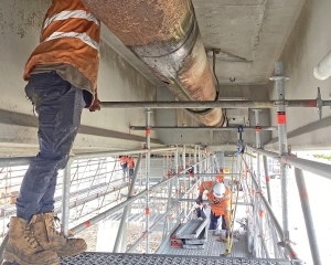 Workers on suspended scaffold replacing brackets, fixtures and fittings on pipes connected to the underside of the San Remo Bridge.